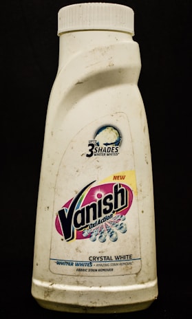 A plastic bottle labeled with the brand Vanish Oxi Action, featuring text promoting its ability to make clothes whiter. The bottle appears somewhat dirty and has a white cap.