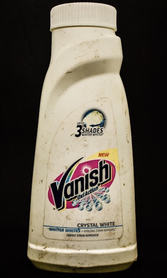 A plastic bottle labeled with the brand Vanish Oxi Action, featuring text promoting its ability to make clothes whiter. The bottle appears somewhat dirty and has a white cap.