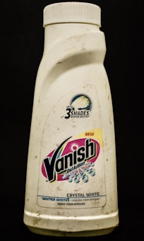 A plastic bottle labeled with the brand Vanish Oxi Action, featuring text promoting its ability to make clothes whiter. The bottle appears somewhat dirty and has a white cap.