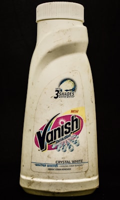 A plastic bottle labeled with the brand Vanish Oxi Action, featuring text promoting its ability to make clothes whiter. The bottle appears somewhat dirty and has a white cap.