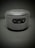A cylindrical electronic speaker with a fabric exterior and a control panel featuring volume buttons and a power button. The color is a dark shade of gray or black, and it is set against a plain, dark background.