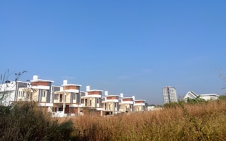 A row of modern, multi-story houses with flat roofs are situated along a grassy field. In the background, a tall apartment building and a few other structures are visible. The sky is clear and blue, indicating a sunny day.