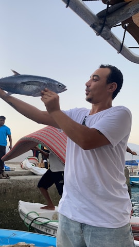 A man in a white t-shirt and jeans holding a fish near the shore. Behind him, a person is tilting a colorful fabric cover, possibly a canopy or tent. The setting appears to be a dock or pier with boats nearby.