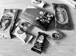 Various packaged snacks are arranged on a wooden surface, including Pocky sticks and multiple KitKat bars. The snacks have Japanese labeling and come in distinctive wrappers featuring almonds, chocolate, and more.