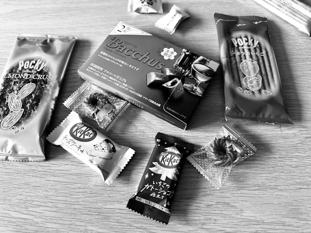 Various packaged snacks are arranged on a wooden surface, including Pocky sticks and multiple KitKat bars. The snacks have Japanese labeling and come in distinctive wrappers featuring almonds, chocolate, and more.