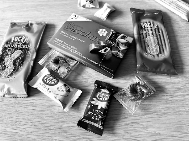 Various packaged snacks are arranged on a wooden surface, including Pocky sticks and multiple KitKat bars. The snacks have Japanese labeling and come in distinctive wrappers featuring almonds, chocolate, and more.