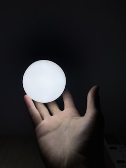 A hand is holding a glowing white orb against a dark background. The soft light from the orb illuminates the hand and casts subtle shadows.