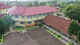 Aerial view of a school complex featuring several buildings with red tiled roofs and light yellow walls. The buildings surround a large paved courtyard with some green planters arranged around its perimeter. A person is standing in the courtyard, and the environment is lush with greenery and trees, suggesting a serene and pleasant atmosphere.