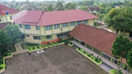 Aerial view of a school complex featuring several buildings with red tiled roofs and light yellow walls. The buildings surround a large paved courtyard with some green planters arranged around its perimeter. A person is standing in the courtyard, and the environment is lush with greenery and trees, suggesting a serene and pleasant atmosphere.