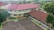 Aerial view of a school complex featuring several buildings with red tiled roofs and light yellow walls. The buildings surround a large paved courtyard with some green planters arranged around its perimeter. A person is standing in the courtyard, and the environment is lush with greenery and trees, suggesting a serene and pleasant atmosphere.