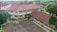 Aerial view of a school complex featuring several buildings with red tiled roofs and light yellow walls. The buildings surround a large paved courtyard with some green planters arranged around its perimeter. A person is standing in the courtyard, and the environment is lush with greenery and trees, suggesting a serene and pleasant atmosphere.