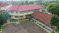 Aerial view of a school complex featuring several buildings with red tiled roofs and light yellow walls. The buildings surround a large paved courtyard with some green planters arranged around its perimeter. A person is standing in the courtyard, and the environment is lush with greenery and trees, suggesting a serene and pleasant atmosphere.