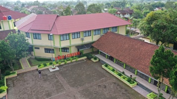 Aerial view of a school complex featuring several buildings with red tiled roofs and light yellow walls. The buildings surround a large paved courtyard with some green planters arranged around its perimeter. A person is standing in the courtyard, and the environment is lush with greenery and trees, suggesting a serene and pleasant atmosphere.