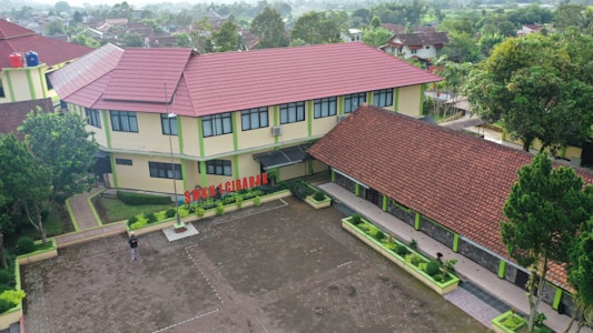 Aerial view of a school complex featuring several buildings with red tiled roofs and light yellow walls. The buildings surround a large paved courtyard with some green planters arranged around its perimeter. A person is standing in the courtyard, and the environment is lush with greenery and trees, suggesting a serene and pleasant atmosphere.