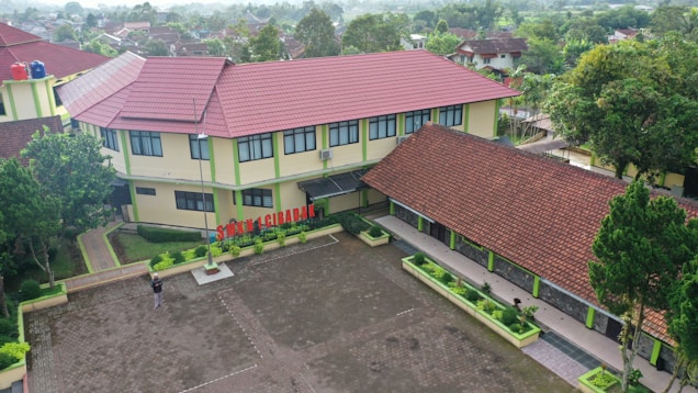 Aerial view of a school complex featuring several buildings with red tiled roofs and light yellow walls. The buildings surround a large paved courtyard with some green planters arranged around its perimeter. A person is standing in the courtyard, and the environment is lush with greenery and trees, suggesting a serene and pleasant atmosphere.