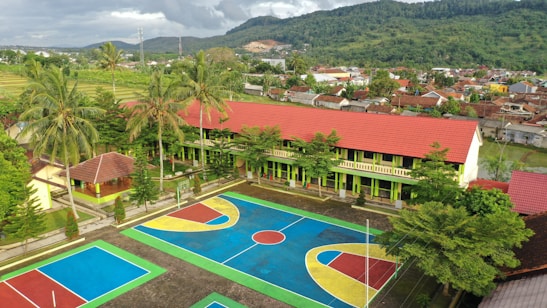 An aerial view of a vibrant school courtyard with a large sports area featuring multiple courts painted in blue, red, yellow, and green. The school building has a long, red roof and is surrounded by palm trees and greenery. In the background, a lush, mountainous landscape is visible alongside a densely packed residential area.