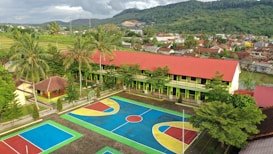 An aerial view of a vibrant school courtyard with a large sports area featuring multiple courts painted in blue, red, yellow, and green. The school building has a long, red roof and is surrounded by palm trees and greenery. In the background, a lush, mountainous landscape is visible alongside a densely packed residential area.