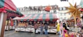 A festive market stall is adorned with colorful striped awnings and garlands, displaying a variety of goods such as candles and decorations. Shoppers, bundled in winter clothing, stand in front of the stall. Decorative stars and plush toys add to the festive atmosphere, with Christmas decorations visible on the roofs.