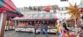 A festive market stall is adorned with colorful striped awnings and garlands, displaying a variety of goods such as candles and decorations. Shoppers, bundled in winter clothing, stand in front of the stall. Decorative stars and plush toys add to the festive atmosphere, with Christmas decorations visible on the roofs.