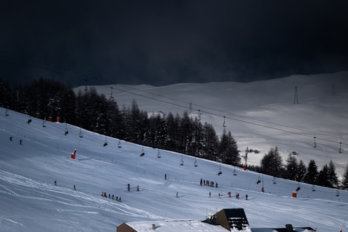 A snow-covered ski slope with people skiing and snowboarding. Trees line the slope, and chairlifts span across the scene, transporting skiers up the hill. The sky appears dark and overcast, creating a contrasting mood against the bright snow.