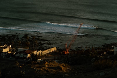 A coastal scene with gentle ocean waves approaching the shore. A construction crane is positioned near the beach, where palm trees line a road adjacent to several small buildings.