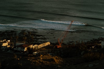 A coastal scene with gentle ocean waves approaching the shore. A construction crane is positioned near the beach, where palm trees line a road adjacent to several small buildings.