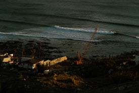 A coastal scene with gentle ocean waves approaching the shore. A construction crane is positioned near the beach, where palm trees line a road adjacent to several small buildings.