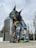 A large lynx sculpture is intricately constructed from colorful recycled materials, such as plastics and metals, forming a vibrant and eye-catching piece of urban art. The background features tall, modern buildings and a row of flags, suggesting a public or city setting.