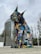 A large lynx sculpture is intricately constructed from colorful recycled materials, such as plastics and metals, forming a vibrant and eye-catching piece of urban art. The background features tall, modern buildings and a row of flags, suggesting a public or city setting.