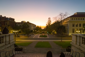 A scenic view of a university campus at sunset, featuring historical buildings, open walkways, and landscaped greenery. Street lights are illuminated as dusk sets in, with students or visitors walking and sitting on the steps in the foreground, observing the tranquil scene.