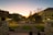 A scenic view of a university campus at sunset, featuring historical buildings, open walkways, and landscaped greenery. Street lights are illuminated as dusk sets in, with students or visitors walking and sitting on the steps in the foreground, observing the tranquil scene.