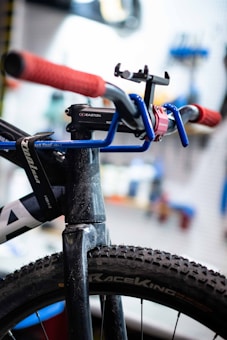 Close-up view of a bicycle featuring a blue handlebar with red grips. The tire is a rugged, all-terrain type and prominently displays the 'RaceKing' branding. A black mount or accessory is attached to the handlebar, and the background is blurred, suggesting a workshop environment.