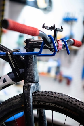 Close-up view of a bicycle featuring a blue handlebar with red grips. The tire is a rugged, all-terrain type and prominently displays the 'RaceKing' branding. A black mount or accessory is attached to the handlebar, and the background is blurred, suggesting a workshop environment.