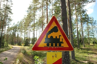 A triangular warning sign with a silhouette of a steam locomotive is prominently displayed in a wooded area. The sign is red and yellow, indicating caution. Tall pine trees and a dirt path are visible in the background under a clear blue sky.