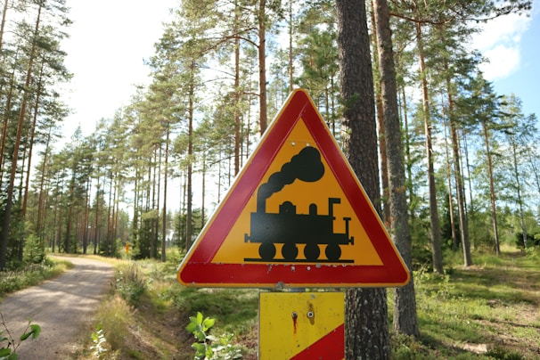 A triangular warning sign with a silhouette of a steam locomotive is prominently displayed in a wooded area. The sign is red and yellow, indicating caution. Tall pine trees and a dirt path are visible in the background under a clear blue sky.