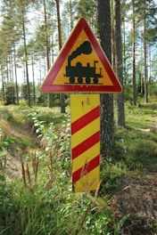 A triangular warning sign featuring a black silhouette of a steam locomotive is mounted on a striped yellow and red post. The sign is placed in a forested area with tall trees and lush greenery, suggesting a rural or natural setting.