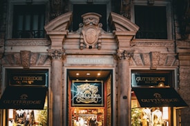 An ornate storefront of a Gutteridge clothing store is seen, featuring classical architectural elements such as decorative columns and intricate moldings. The display windows are illuminated and exhibit mannequins dressed in elegant suits. The store signage is prominently displayed above the entrance, with rich wood and glass doors enhancing the sophisticated ambiance.