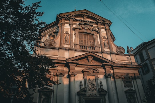 The image features an ornate, historical building likely a church or cathedral with classical architectural details including columns, sculptures, and intricate stone carvings. The facade is illuminated by warm sunlight which accentuates its textures and details. Tall trees partially obscure the building on the left.