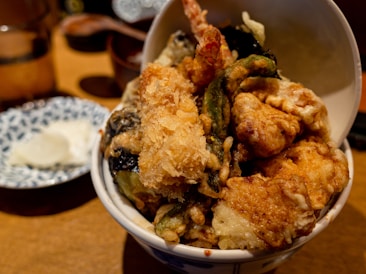 A bowl of assorted tempura, featuring deep-fried shrimp, vegetables, and seafood. The tempura is golden and crispy, served atop a bowl of rice. In the background, there is a small plate with a white dipping sauce.