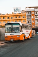 A vintage orange and white bus is parked on a street in front of a yellow hotel building, with a sign on top that reads 'Sea Garden Hurghada Red Sea.' The hotel has balconies and several satellite dishes on the roof. A modern building is adjacent to the hotel.