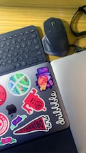 A workspace featuring a wireless keyboard, a mouse, and a pair of eyeglasses on a wooden surface. A laptop adorned with colorful stickers, including abstract designs and typography, adds a vibrant touch.