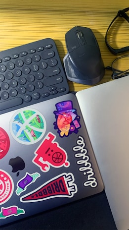 A workspace featuring a wireless keyboard, a mouse, and a pair of eyeglasses on a wooden surface. A laptop adorned with colorful stickers, including abstract designs and typography, adds a vibrant touch.