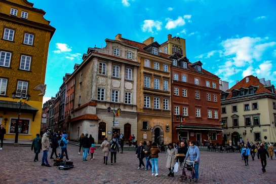 A European city square with historical, colorful buildings featuring various architectural styles. Many people are walking or standing around the square, some with shopping bags, enjoying the clear day under a bright blue sky with scattered clouds.