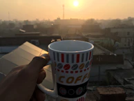 A hand holding a coffee mug with the gold butterfly logo, sunlight casting a warm glow.