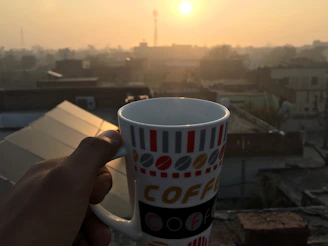 A hand holding a coffee mug with the gold butterfly logo, sunlight casting a warm glow.