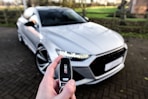 A person holds a car key fob with a focus on an RS logo, in the foreground. In the background, a white, sporty, luxury car is parked on a paved driveway. The scene is outdoors with a blurred view of a garden or park area with green grass and trees.