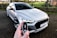 A person holds a car key fob with a focus on an RS logo, in the foreground. In the background, a white, sporty, luxury car is parked on a paved driveway. The scene is outdoors with a blurred view of a garden or park area with green grass and trees.