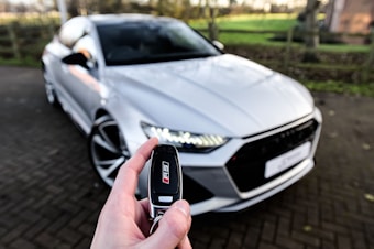 A person holds a car key fob with a focus on an RS logo, in the foreground. In the background, a white, sporty, luxury car is parked on a paved driveway. The scene is outdoors with a blurred view of a garden or park area with green grass and trees.