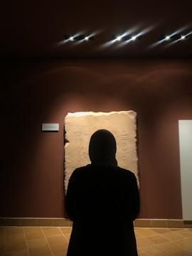 A person stands in front of an ancient stone tablet with inscriptions, which is displayed on a wall under focused lighting. The person's silhouette is highlighted against the lit artifact, creating a contemplative atmosphere.