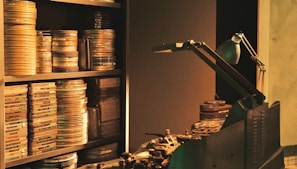 Film reels are stacked on shelves and a table, alongside a vintage film editing machine. A desk lamp provides dim, focused light over the area.